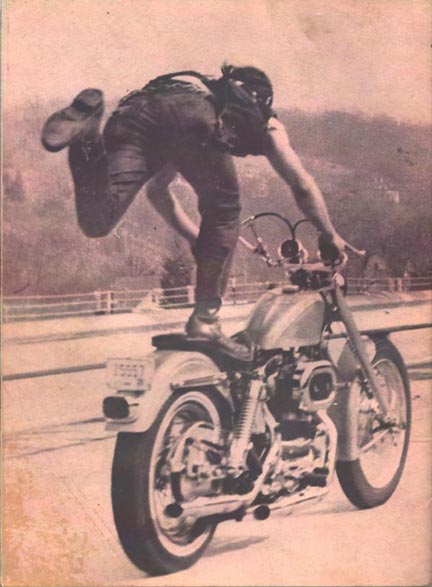 Vintage photo of a biker standing on the seat of a motorcyle with one leg in the air.